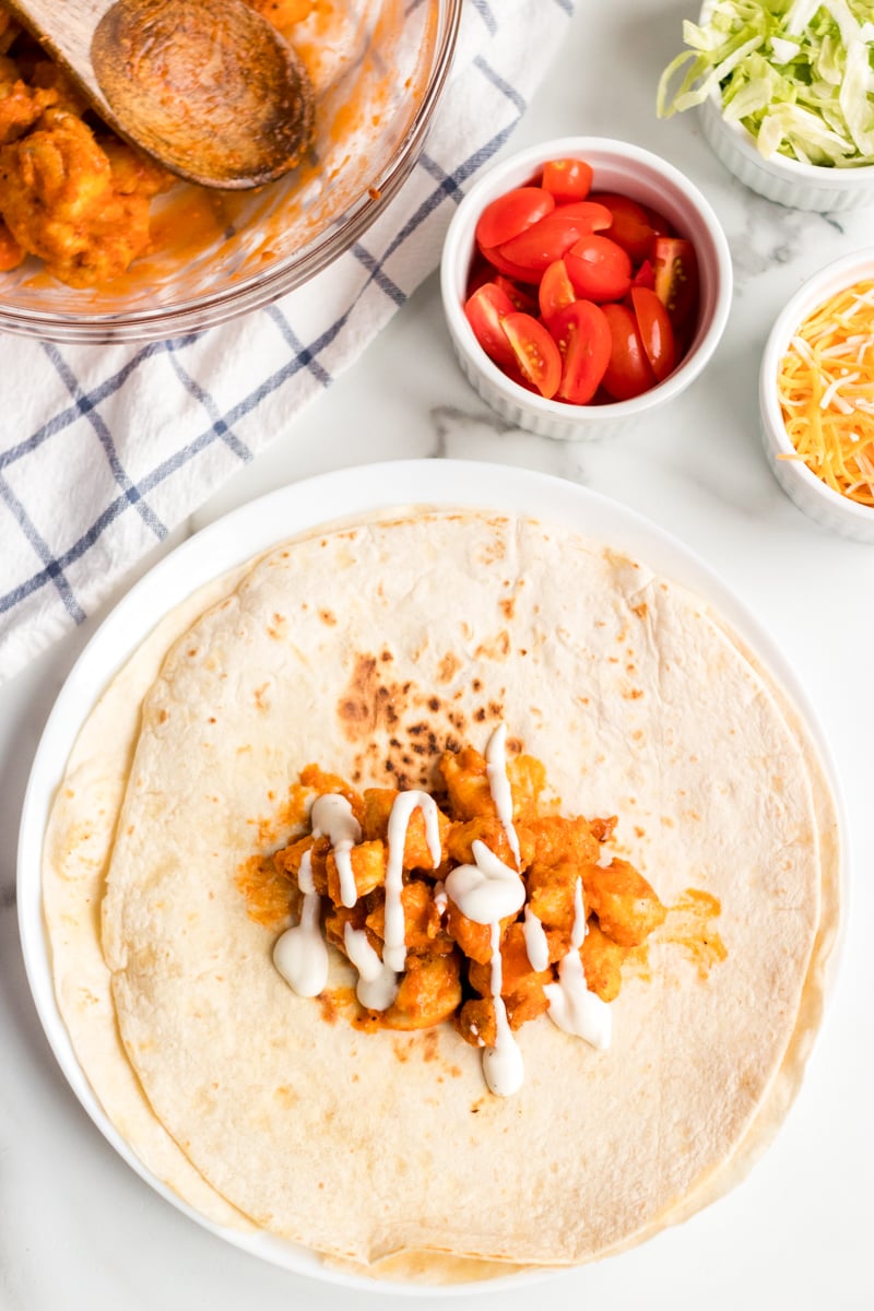 If you like the Buffalo Ranch Chicken Wrap from Buffalo Wild Wings, you'll love this lightened up homemade version! Lightly breaded buffalo chicken pieces wrapped up in a soft tortilla with ranch dressing, shredded lettuce, cheese, and tomatoes make a great dinner or the perfect game day meal! | www.persnicketyplates.com buffalo chicken on a flour tortilla for buffalo ranch chicken wraps