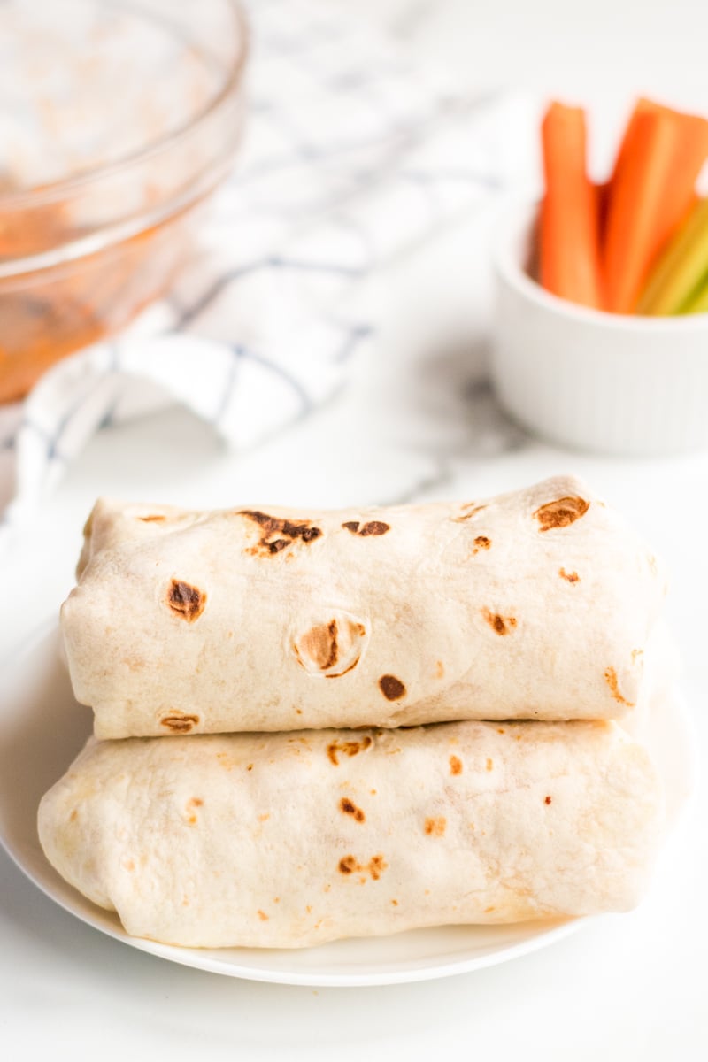 If you like the Buffalo Ranch Chicken Wrap from Buffalo Wild Wings, you'll love this lightened up homemade version! Lightly breaded buffalo chicken pieces wrapped up in a soft tortilla with ranch dressing, shredded lettuce, cheese, and tomatoes make a great dinner or the perfect game day meal! | www.persnicketyplates.com wrapped buffalo ranch chicken wraps stacked with carrots in background