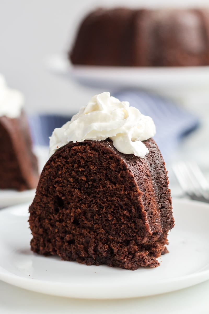 This rich, dense Sour Cream Chocolate Bundt Cake is a copycat of the famous Williams Sonoma recipe. Perfect for those chocolate cravings! | www.persnicketyplates.com single slice of sour cream chocolate bundt cake on white plate topped with whipped cream