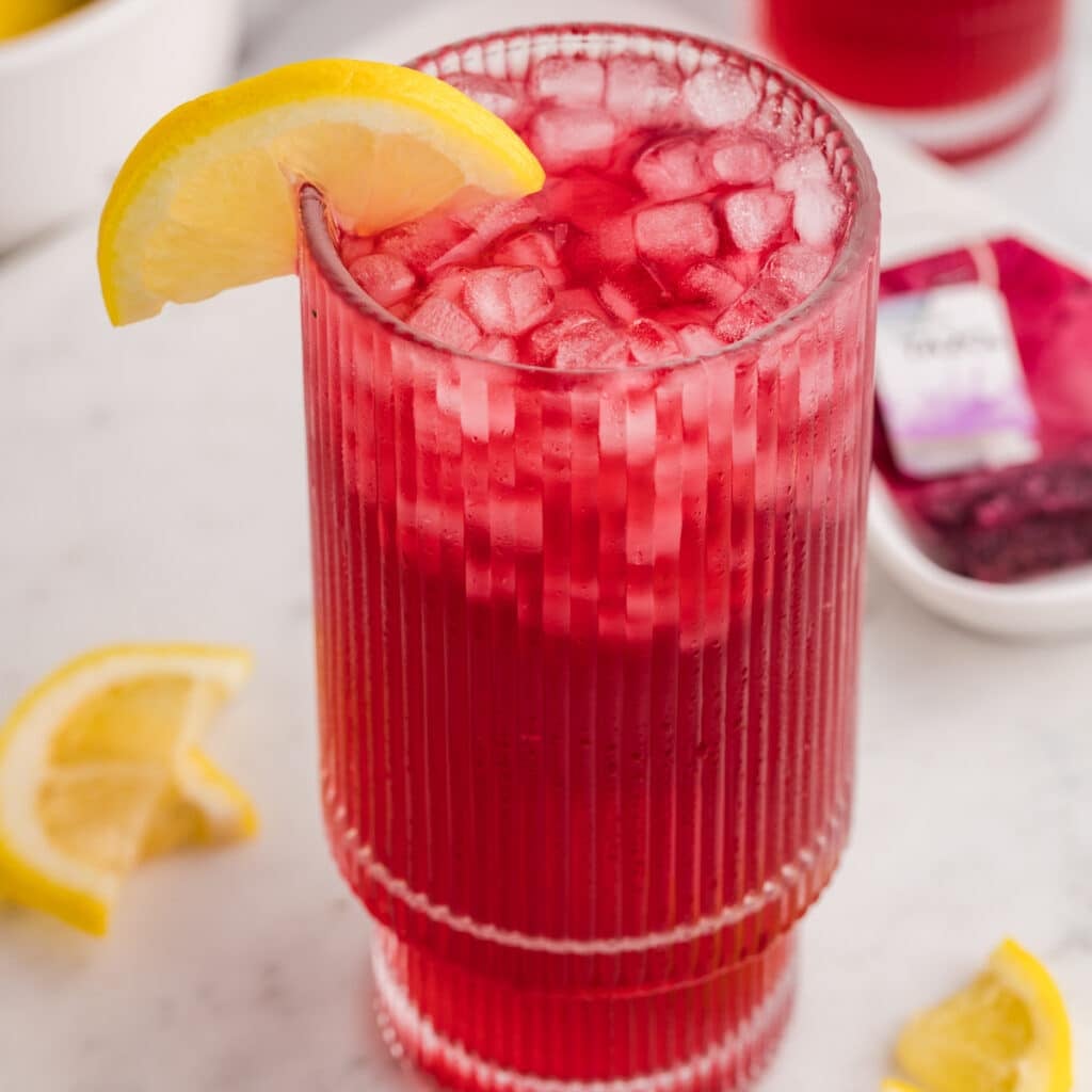 A tall ribbed glass filled with pink iced tea and a lemon wedge.