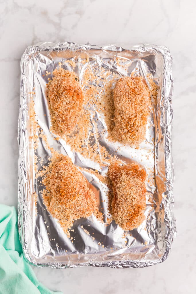 Four breaded stuffed chicken breasts on a foil-lined baking sheet.