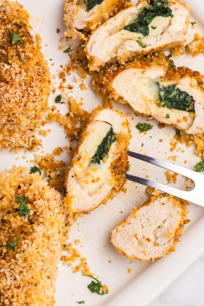 A close-up of a crispy, golden-breaded Cajun stuffed chicken breast sliced open to reveal melted cheese and spinach filling, with crumbs scattered on a white plate and a serving fork lifting one piece.