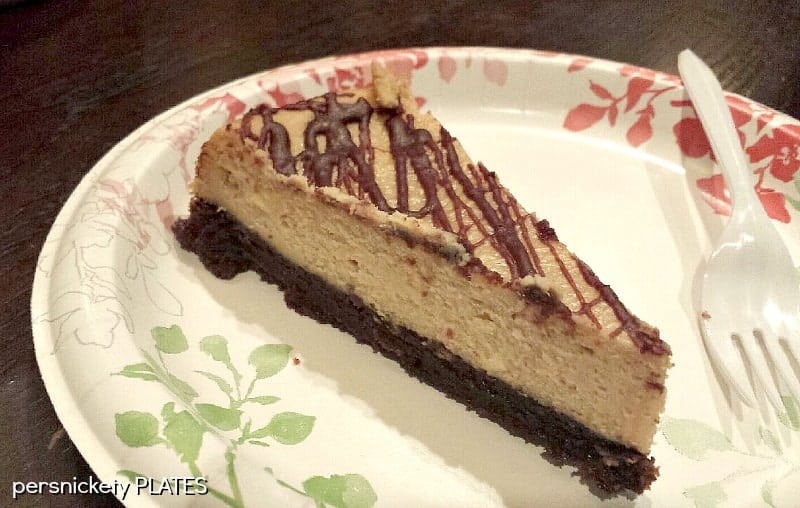 slice of Brownie Bottom Peanut Butter Cheesecake on paper plate Brownie Bottom Peanut Butter Cheesecake has a layer of chewy brownie topped with a creamy peanut butter cheesecake and drizzled with rich chocolate. | www.persnicketyplates.com #cheesecake #peanutbutter #brownie #dessert
