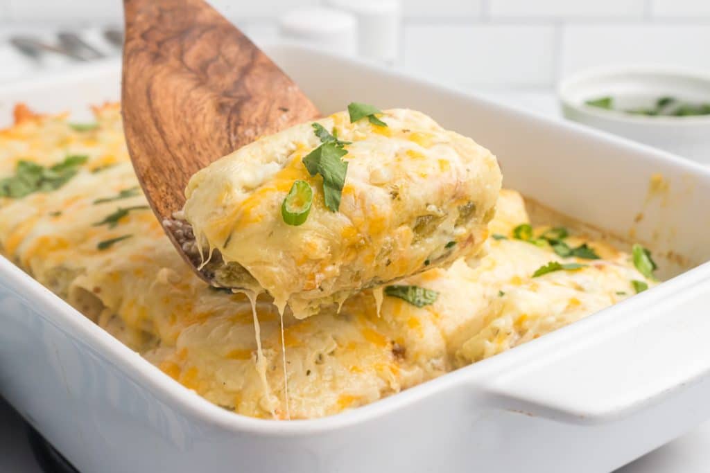 wooden spoon lifting a green enchilada from a baking dish.