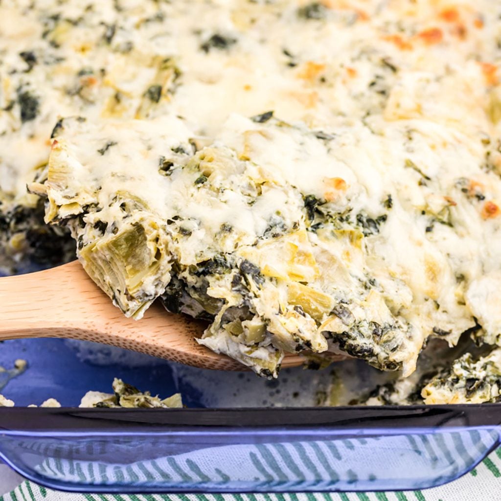 wooden spoon scooping spinach artichoke chicken casserole from a blue glass dish.