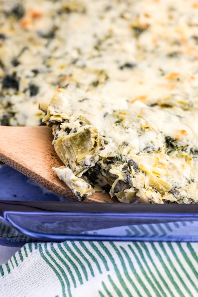 wooden spoon scooping spinach dip casserole from a blue baking dish.