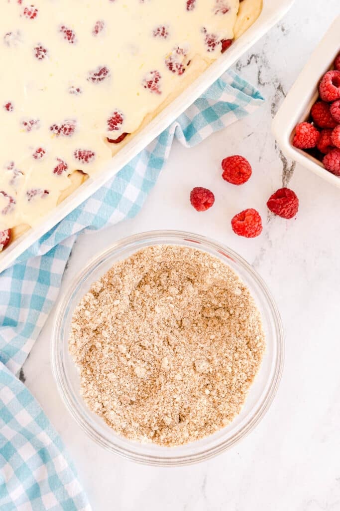 Glass bowl of crumb topping next to a pan of batter with raspberries and a container of raspberries on the side.
