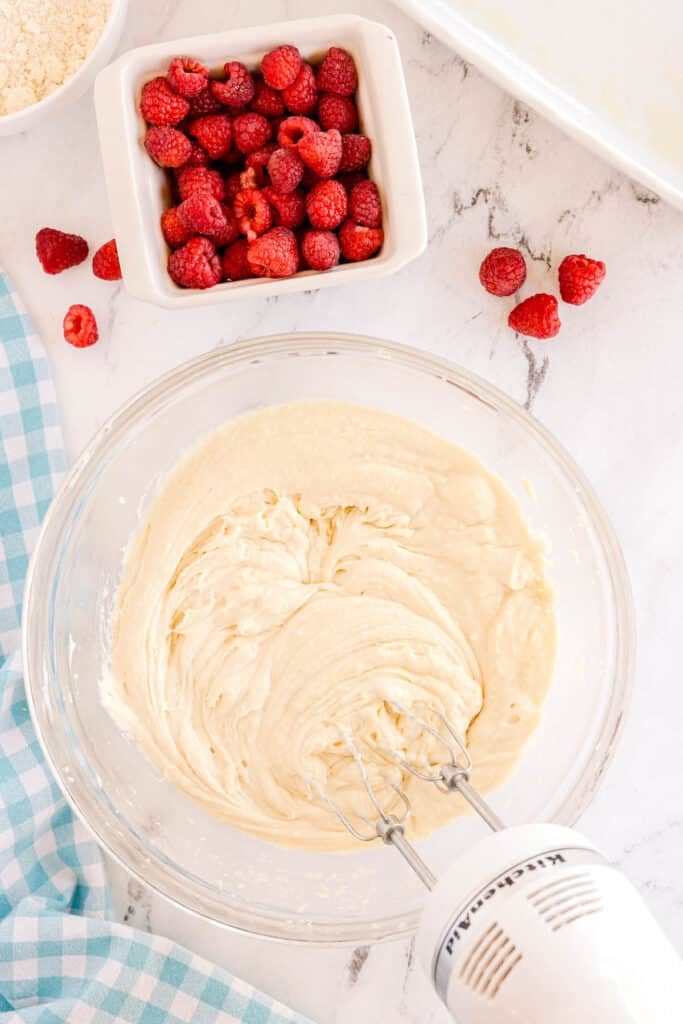 cake batter in a mixing bowl with a hand mixer surrounded by raspeberries.