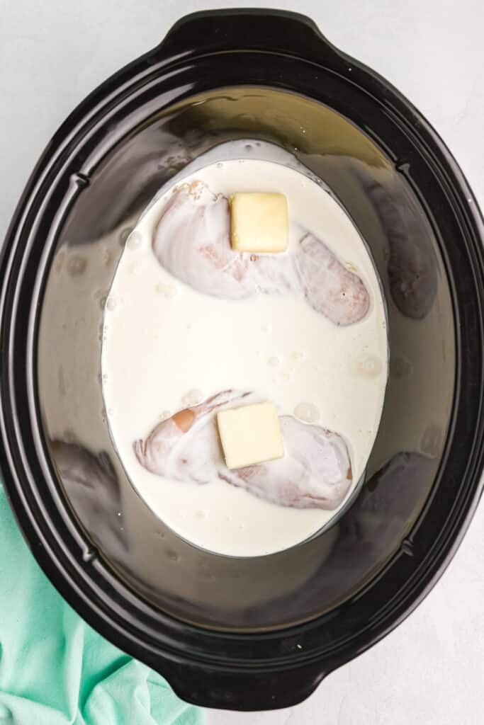 overhead shot of raw chicken breasts, butter, and cream in a slow cooker.