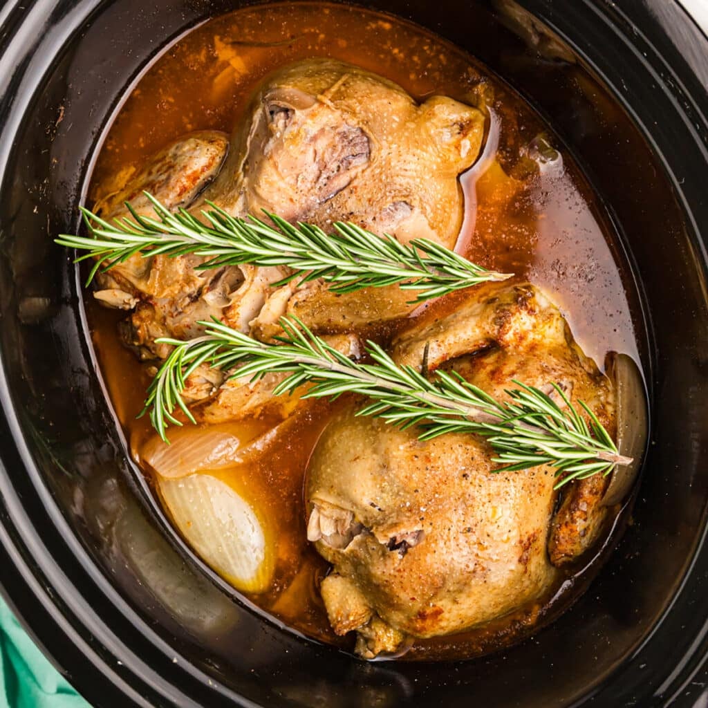 Two cooked Cornish hens in a crockpot with sprigs of rosemary.