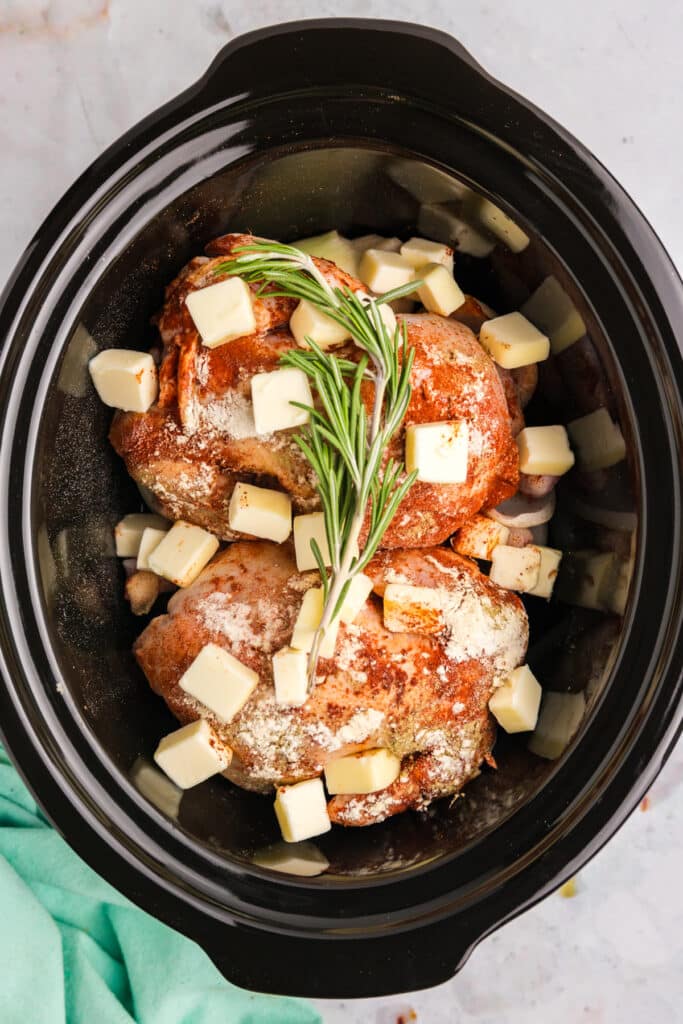 Two raw Cornish hens in a crockpot, seasoned and topped with small butter cubes and sprigs of fresh rosemary, ready to slow cook.