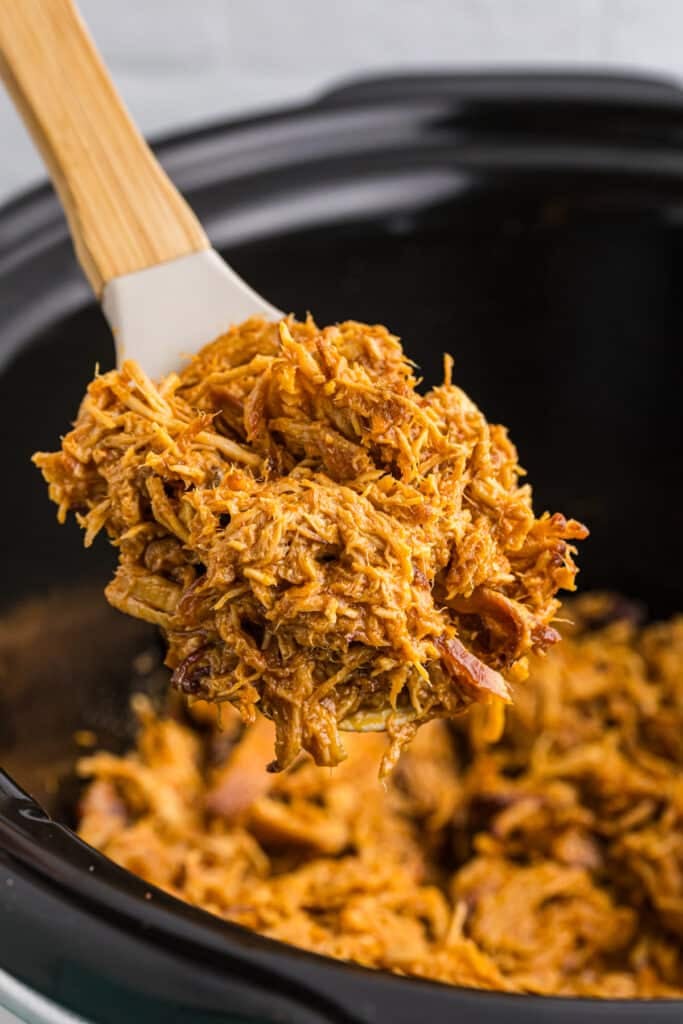 spoon lifting a scoop of shredded hot honey chicken from a slow cooker.
