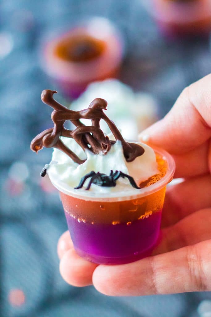 hand holding a jello shot topped with whipped cream, a chocolate cobweb, and a plastic spider.
