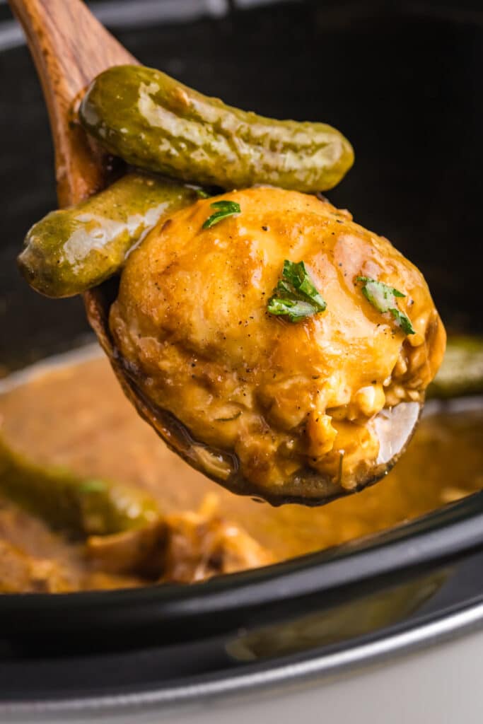 spoon lifting a chicken thigh with pickles from a crockpot.