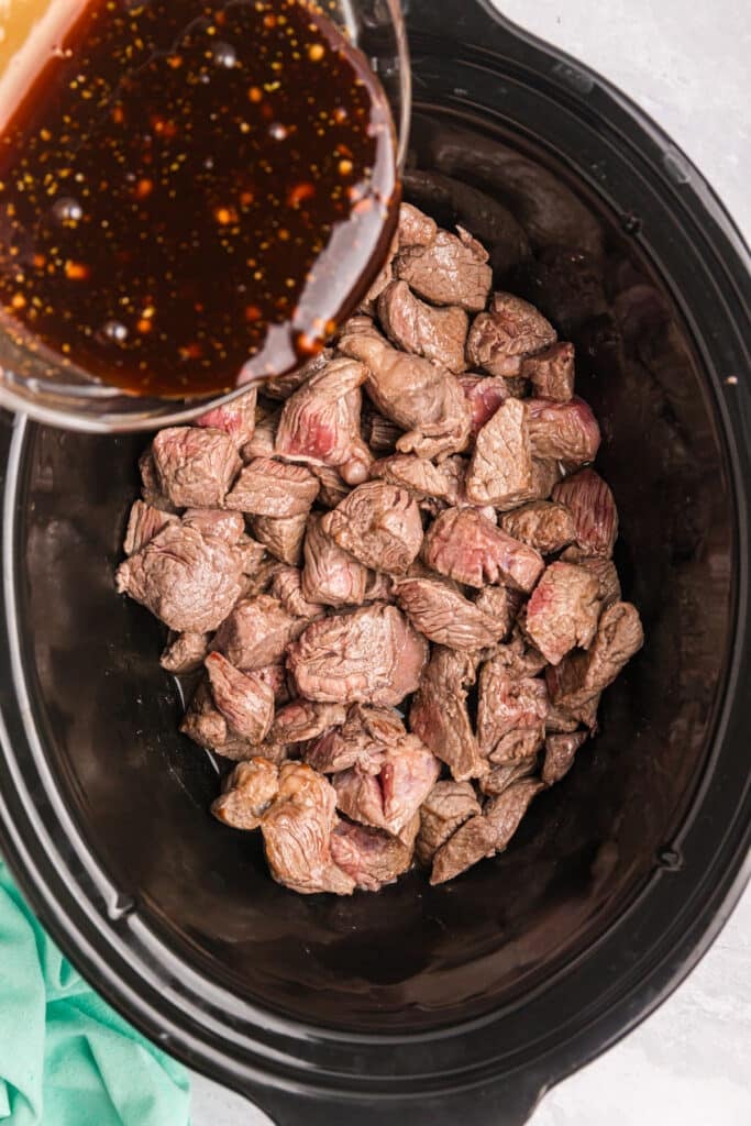 sauce pouring onto seared beef chunks in a crockpot.