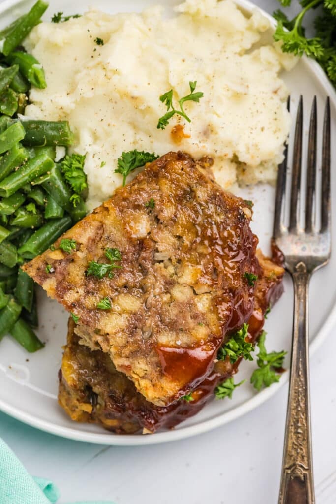 slice of meatloaf on a plate with mashed potatoes & green beans.