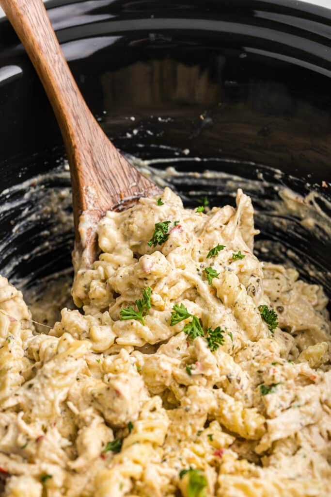 wooden spoon scooping chicken pasta from a slow cooker.