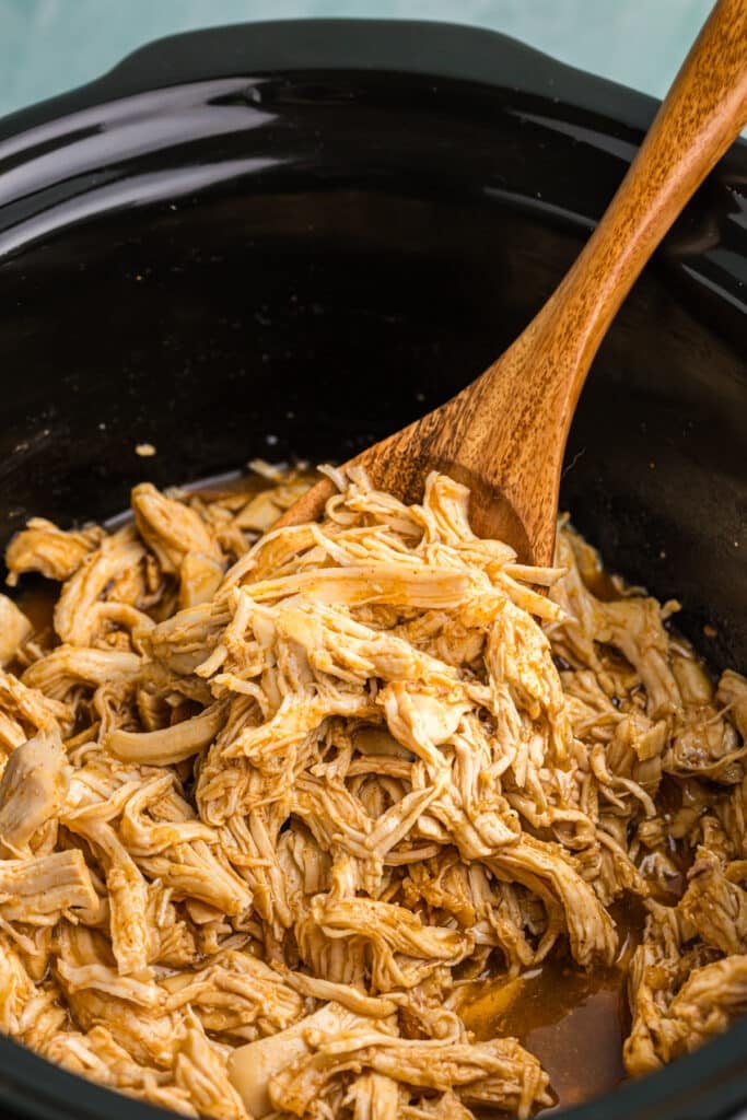 wooden spoon lifting a scoop of shredded chicken from a slow cooker.