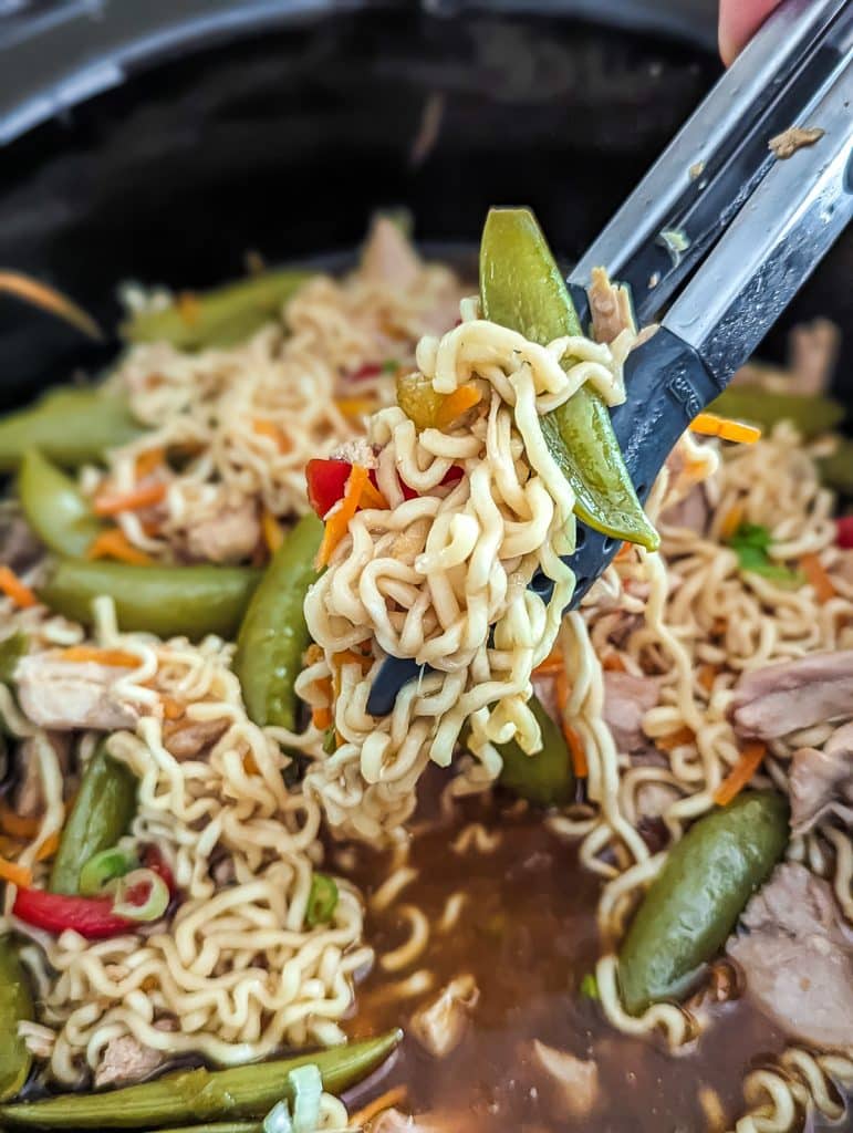 tongs lifting chicken ramen from a crockpot.