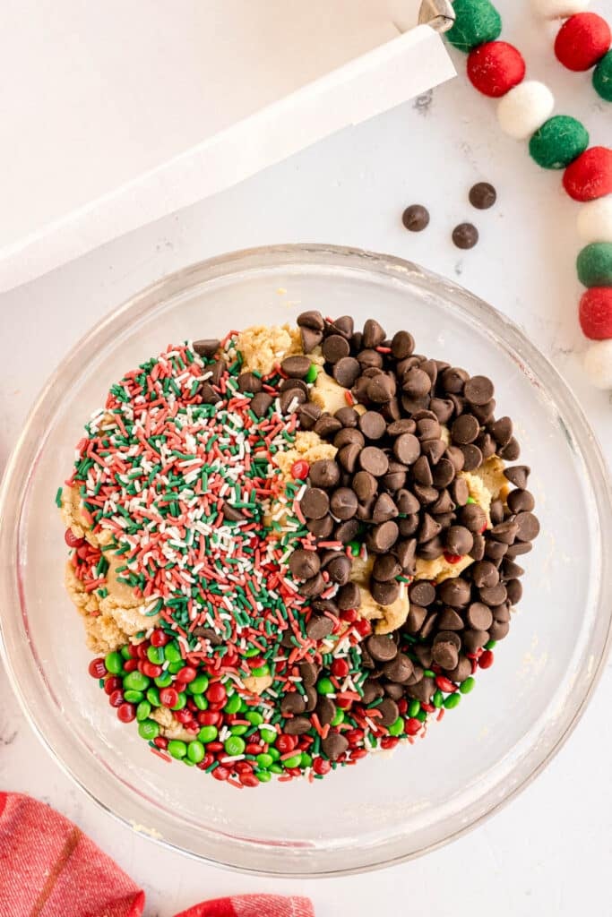 m&ms, chocolate chips, christmas sprinkles in a mixing bowl with cookie dough.