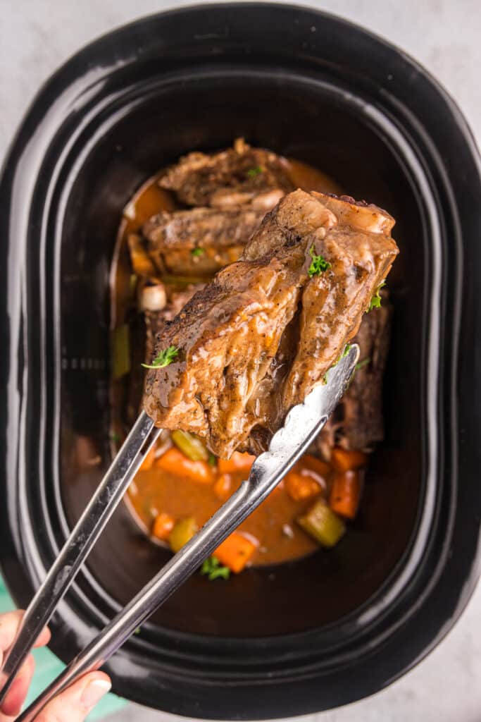tongs lifting short ribs from a crockpot.