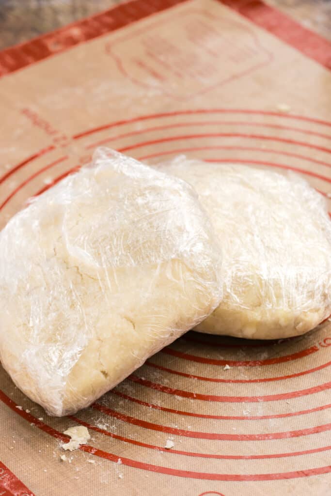 two squares of pie dough wrapped in plastic wrap on a silicone mat.