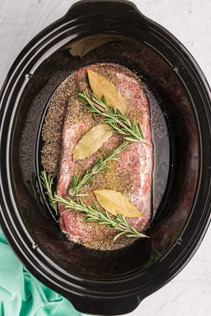a pork loin topped with rosemary and bay leaves in a crockpot.