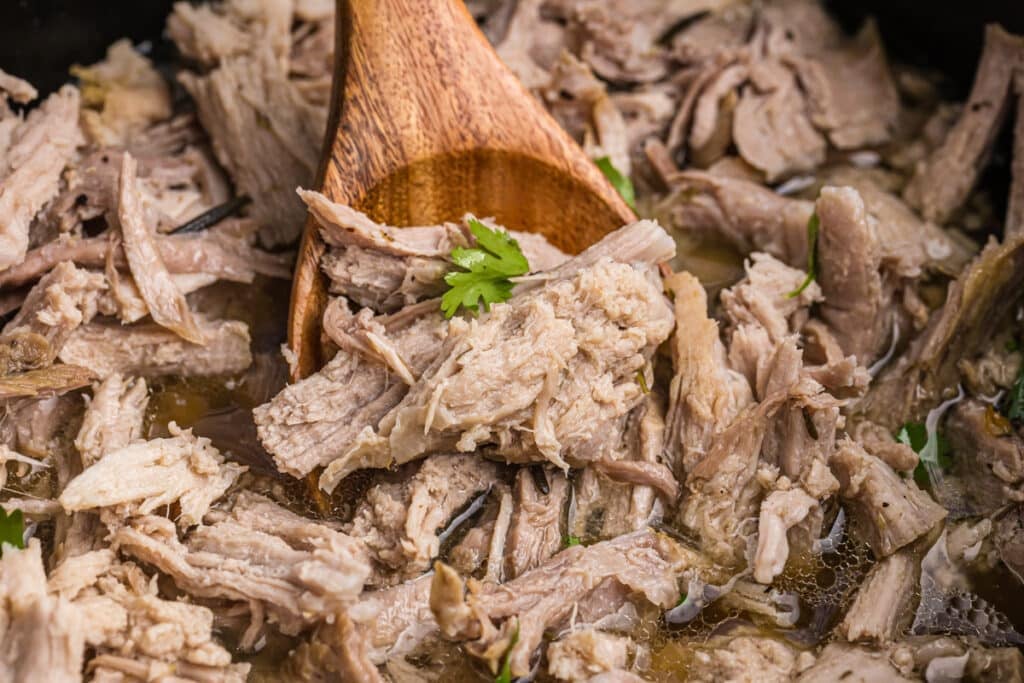 close up of a wooden spoon lifting a scoop of carnitas from a slow cooker.