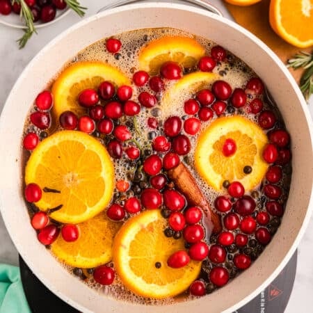 A pot with orange slices, cranberries, rosemary, and cinnamon sticks in liquid.