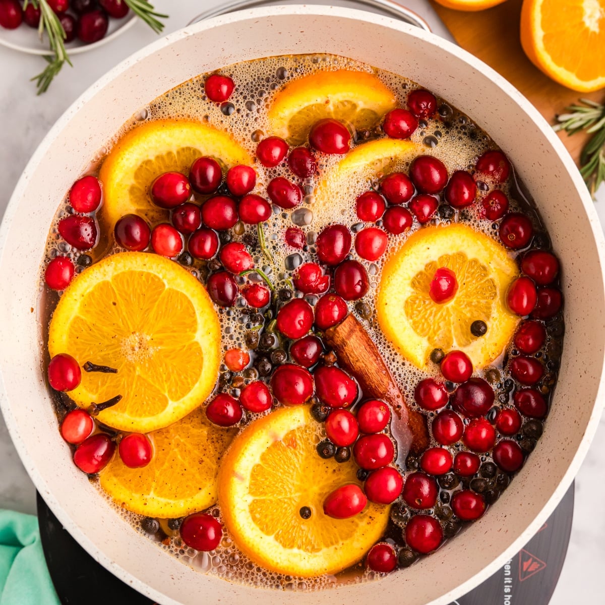 A pot with orange slices, cranberries, rosemary, and cinnamon sticks in liquid.