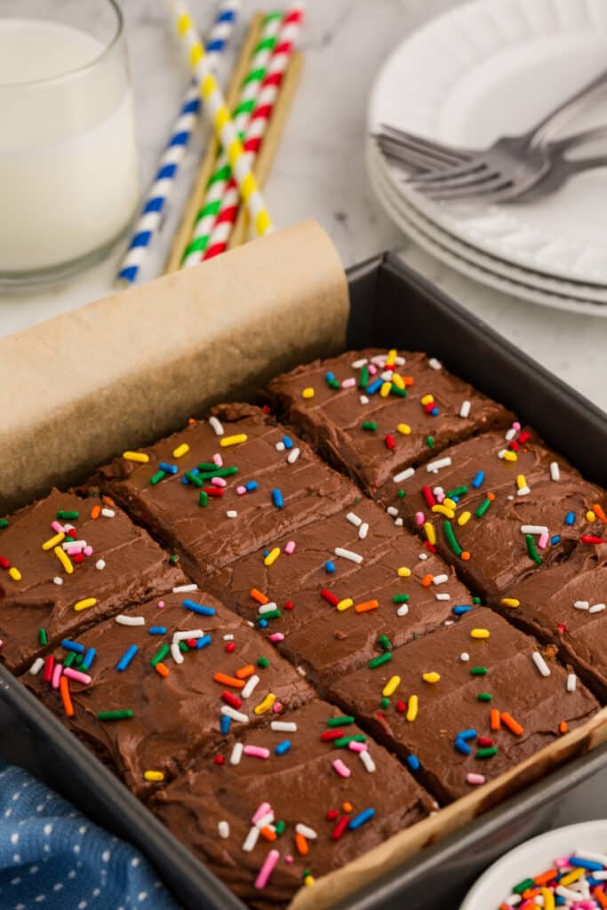 A pan of confetti snack cake with chocolate frosting and rainbow sprinkles, sliced into squares, with colorful straws and a glass of milk in the background.