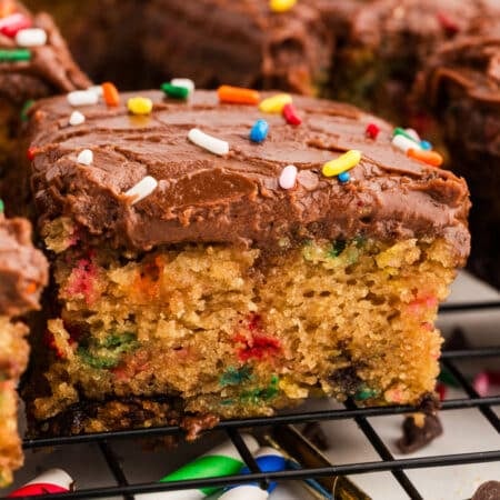 Close-up of a slice of confetti cake with colorful sprinkles in the cake and topped with chocolate frosting and rainbow sprinkles.