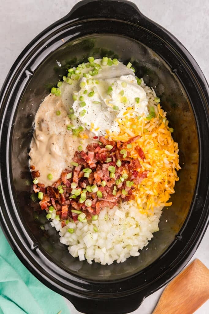 A top-down view of a slow cooker containing shredded hash browns, chopped onions, shredded cheese, sour cream, cream of onion soup, cooked bacon, and scallions before mixing.