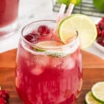 A glass of cranberry punch with lime slice, cranberries, and pink-striped straws on a wooden tray.