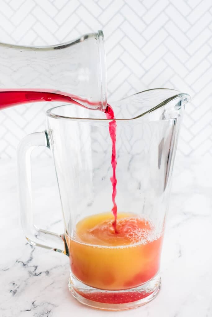 Cranberry juice being poured into a glass pitcher with pineapple juice.