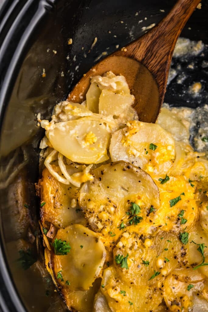 A wooden spoon scooping scalloped potatoes from a crockpot.