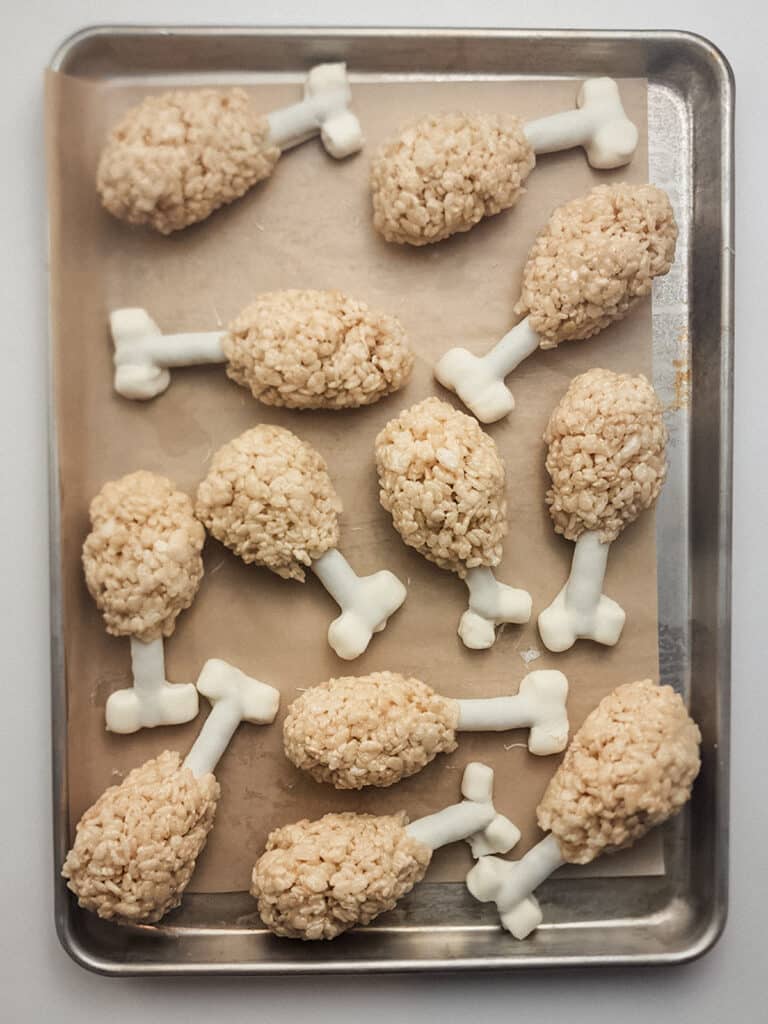 A tray of Rice Krispies treats shaped like turkey legs with white candy bones on parchment paper.