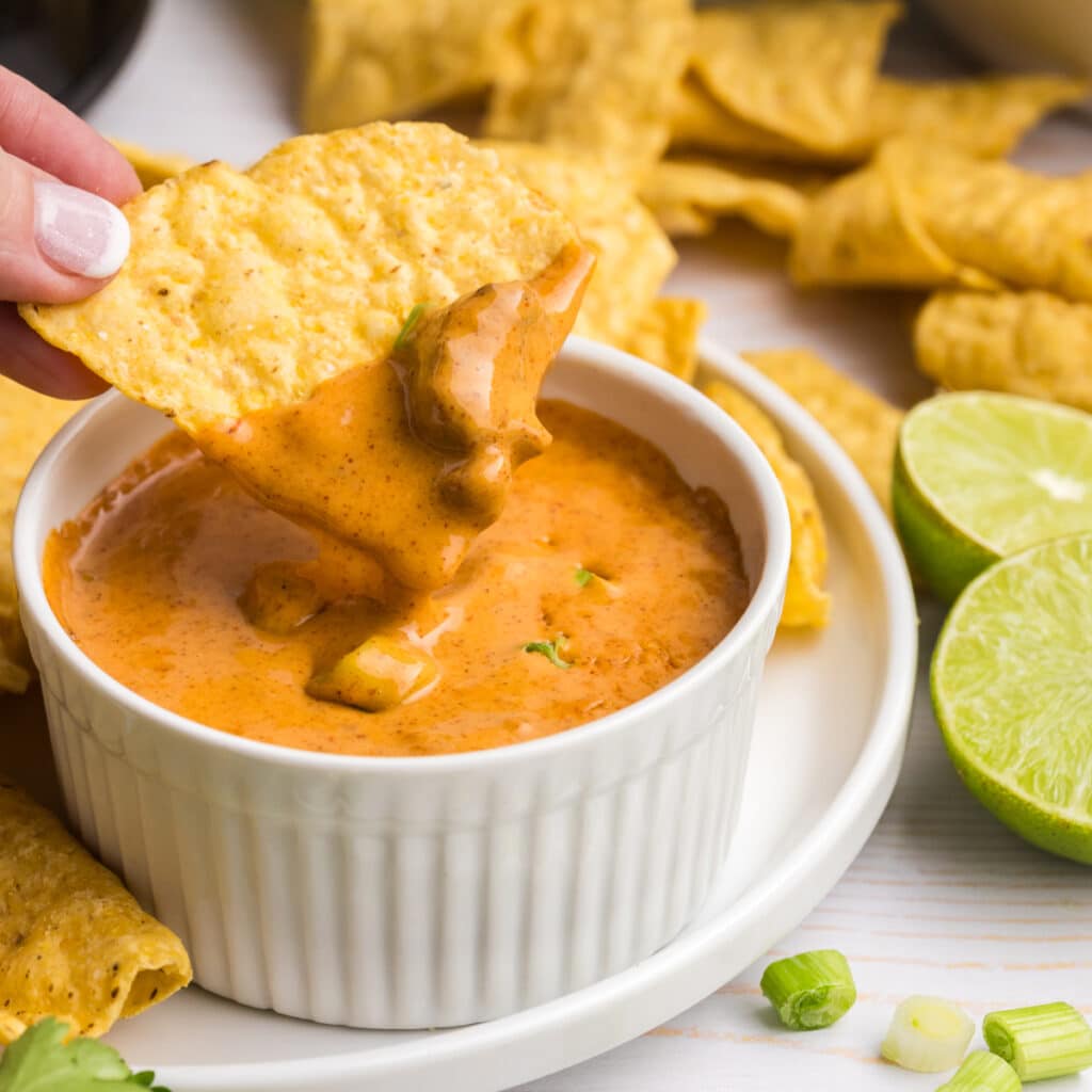 A tortilla chip dipped into queso in a white ramekin with lime halves and chips around.