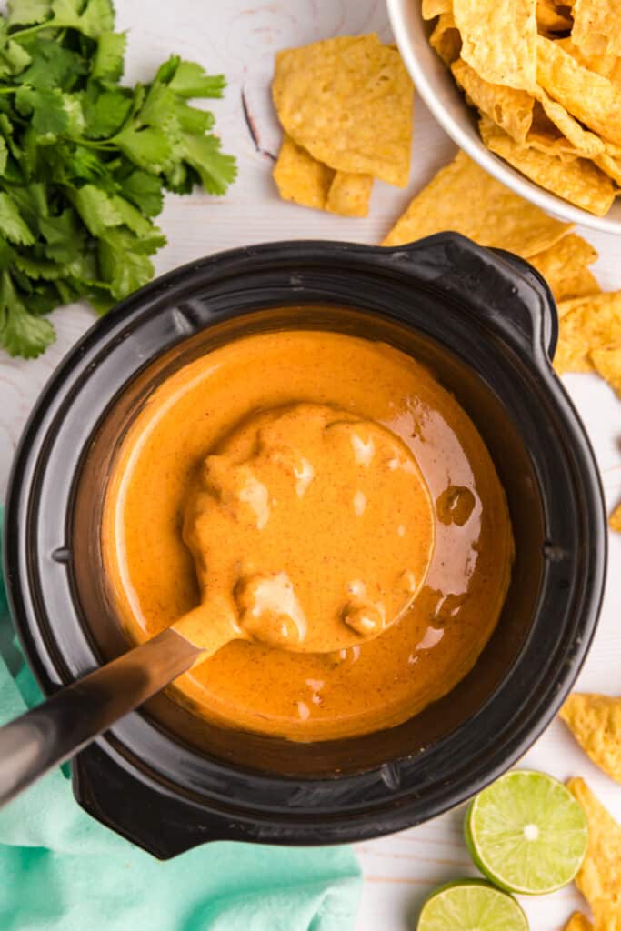 A ladle of creamy queso dip in a crockpot, surrounded by limes and chips.