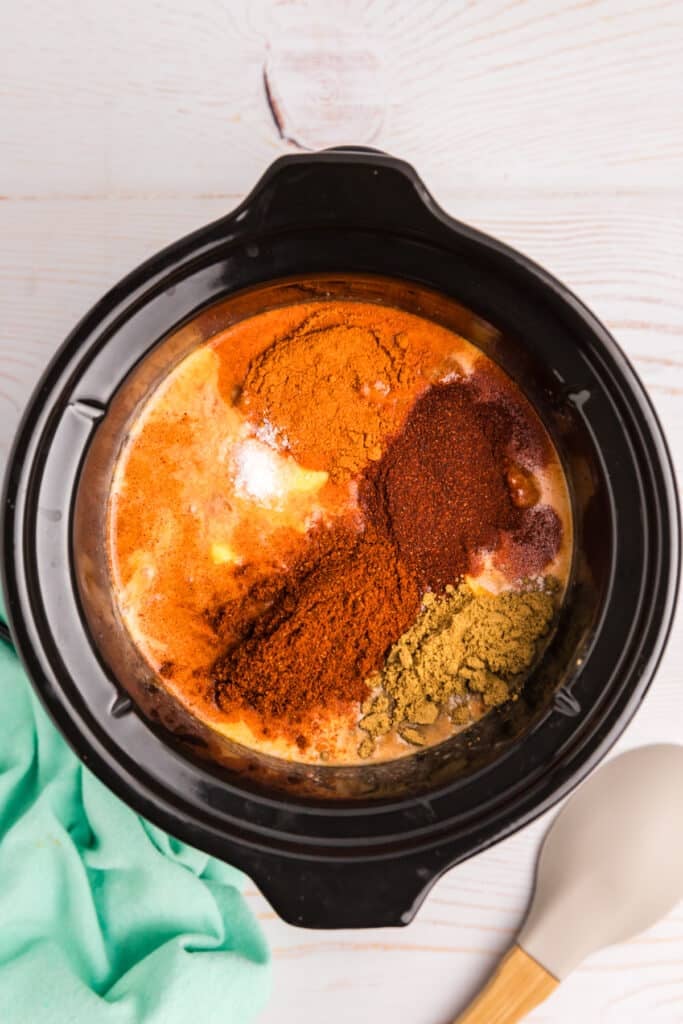A crockpot filled with layered ingredients for queso, including spices, cheese, and chili.