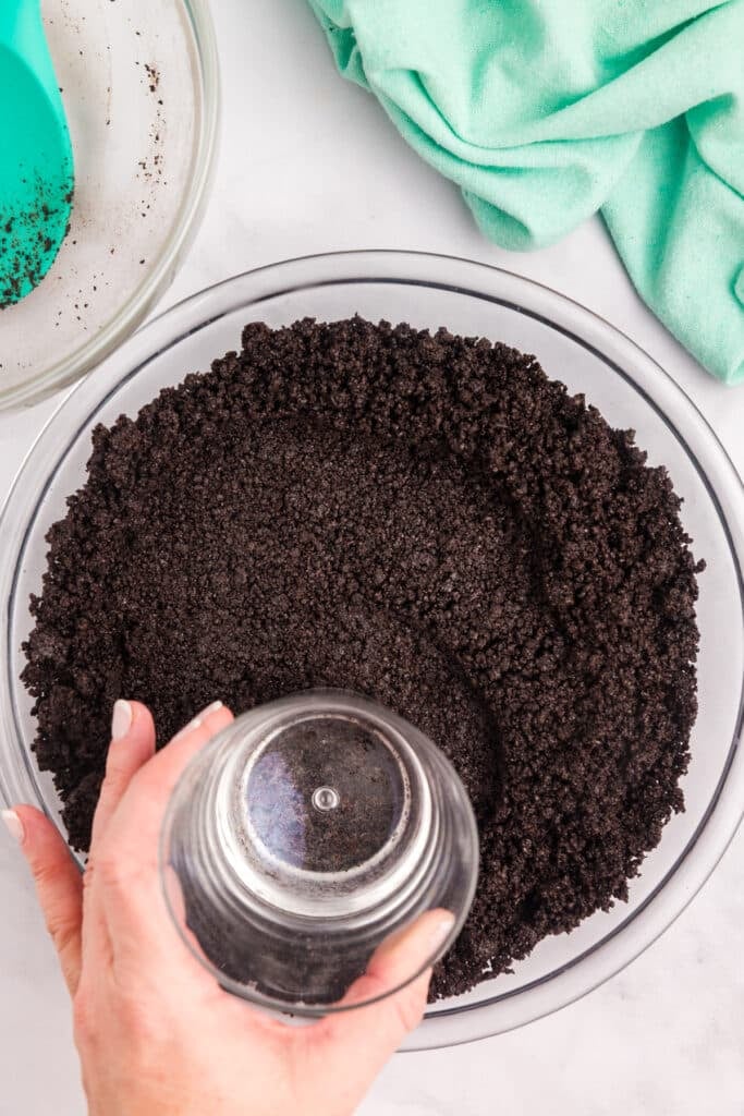 A hand using a glass cup to press Oreo crumbs gently into the sides and bottom of a glass pie plate to form a crust.