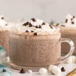 A glass mug filled with hot chocolate topped with whipped cream, mini chocolate chips, and marshmallows.