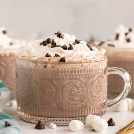 A glass mug filled with hot chocolate topped with whipped cream, mini chocolate chips, and marshmallows.