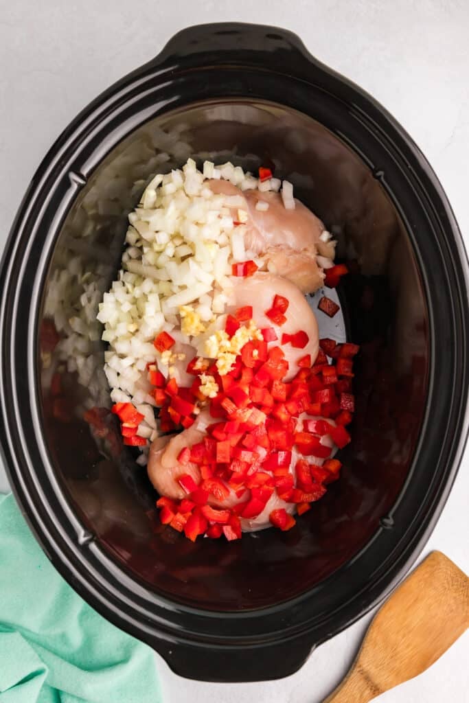 overhead shot of chicken breasts, diced peppers, onions in a black crockpot.