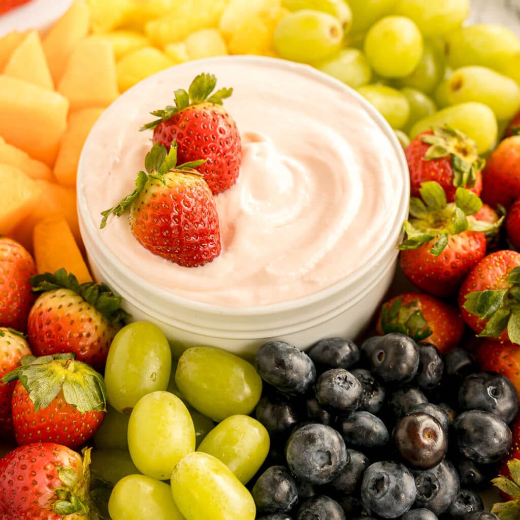 A bowl of creamy fruit dip surrounded by fresh strawberries, grapes, blueberries, and melon.