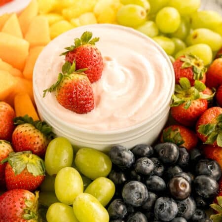 A bowl of creamy fruit dip surrounded by fresh strawberries, grapes, blueberries, and melon.