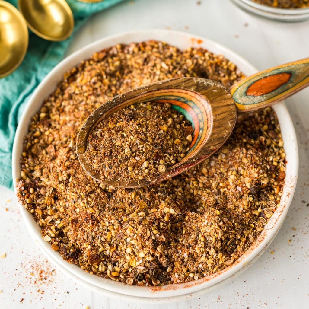 A bowl of homemade Montreal steak seasoning with a rustic wooden spoon scooping some up.