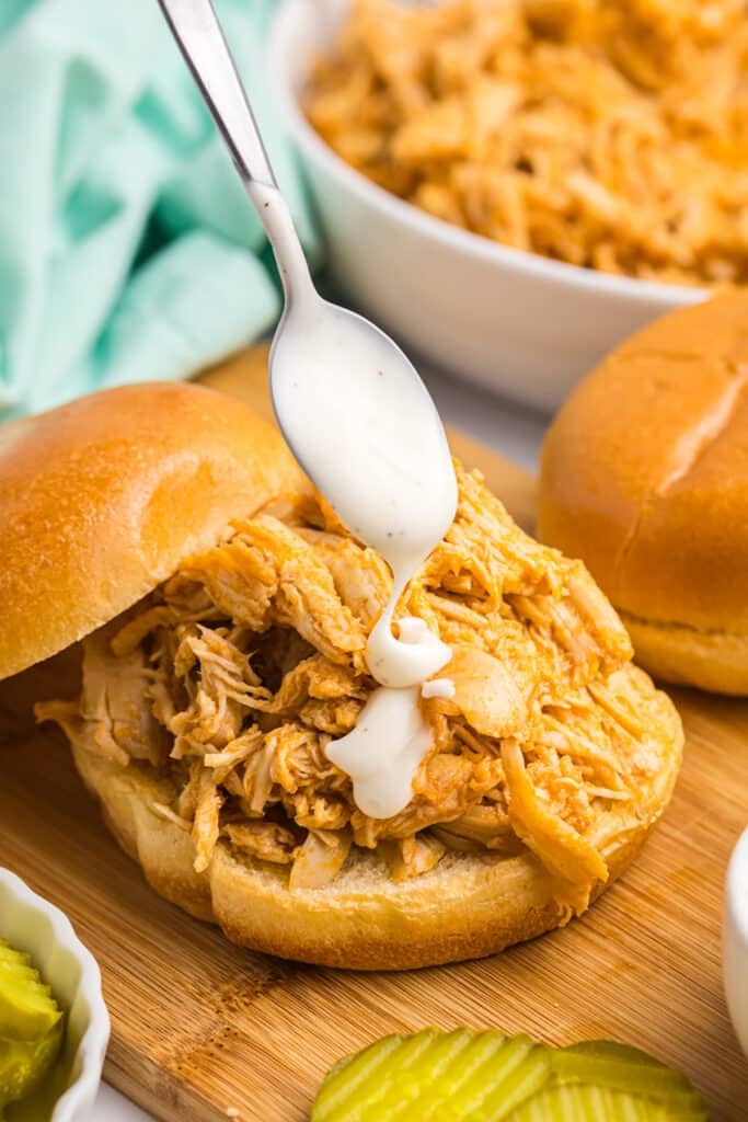 spoon drizzling ranch dressing over a shredded chicken sandwich.