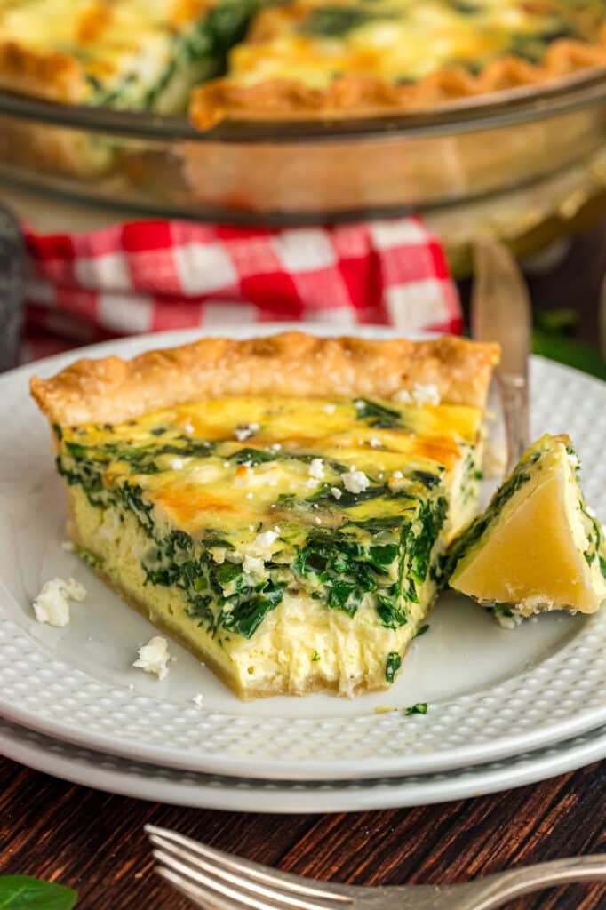 A slice of spinach feta quiche served on a white plate with a fork, showing the fluffy egg filling and crust.