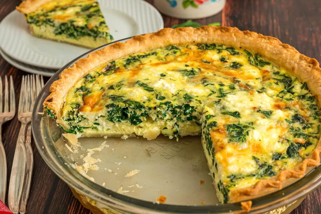 A partially sliced spinach and cheese quiche in a glass pie dish, with a plated slice in the background.